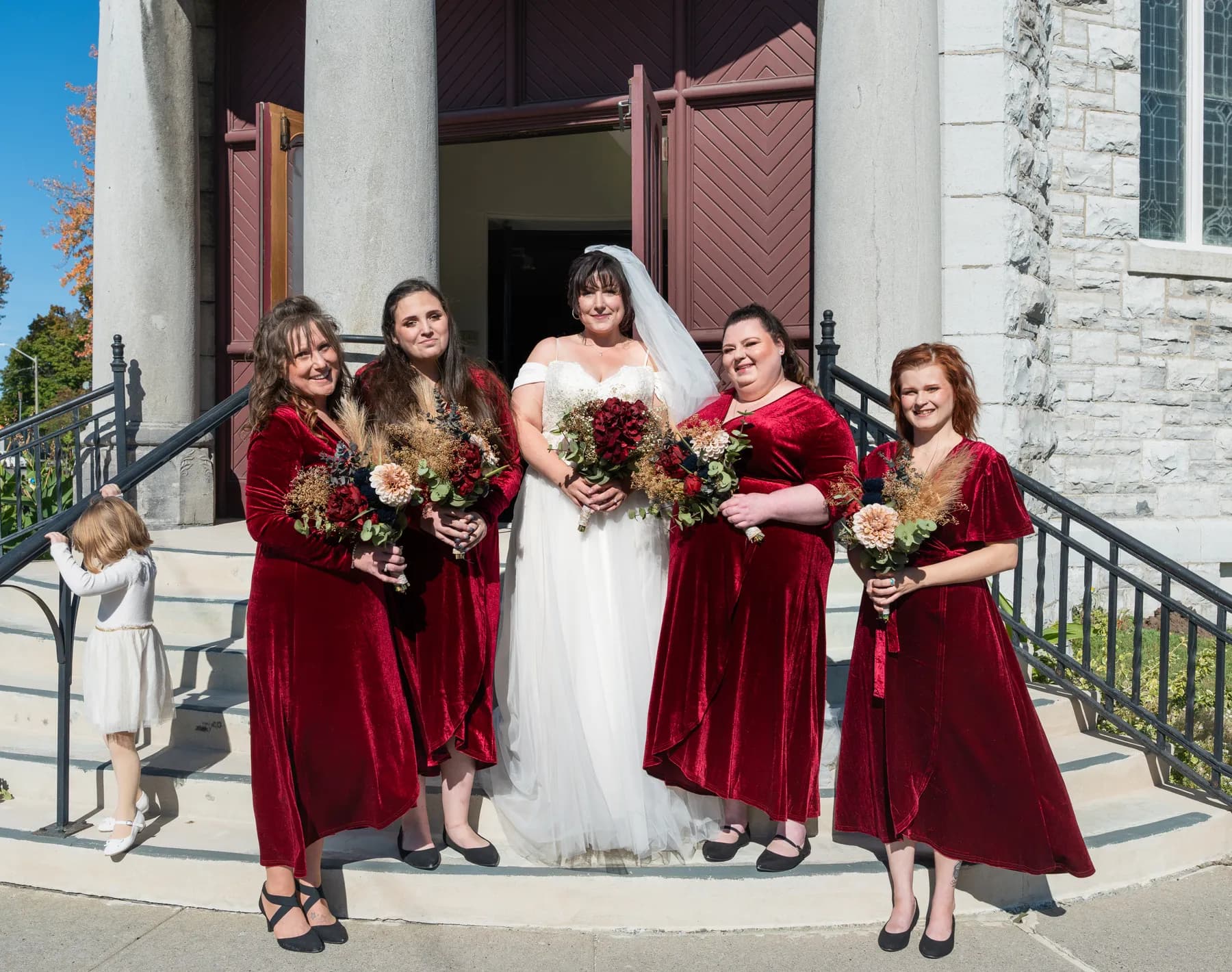 The bride with bridesmaids and a flower girl outside the church at Kody and Jaz’s wedding.