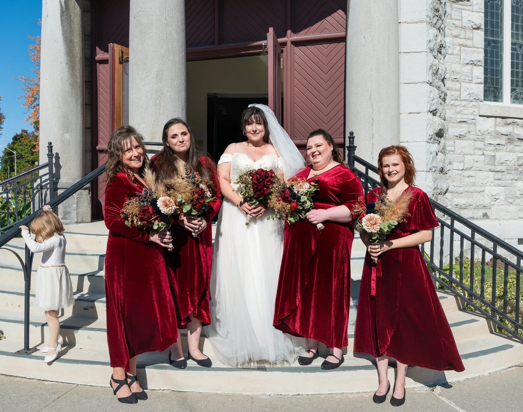 The bride with bridesmaids and a flower girl outside the church at Kody and Jaz’s wedding.