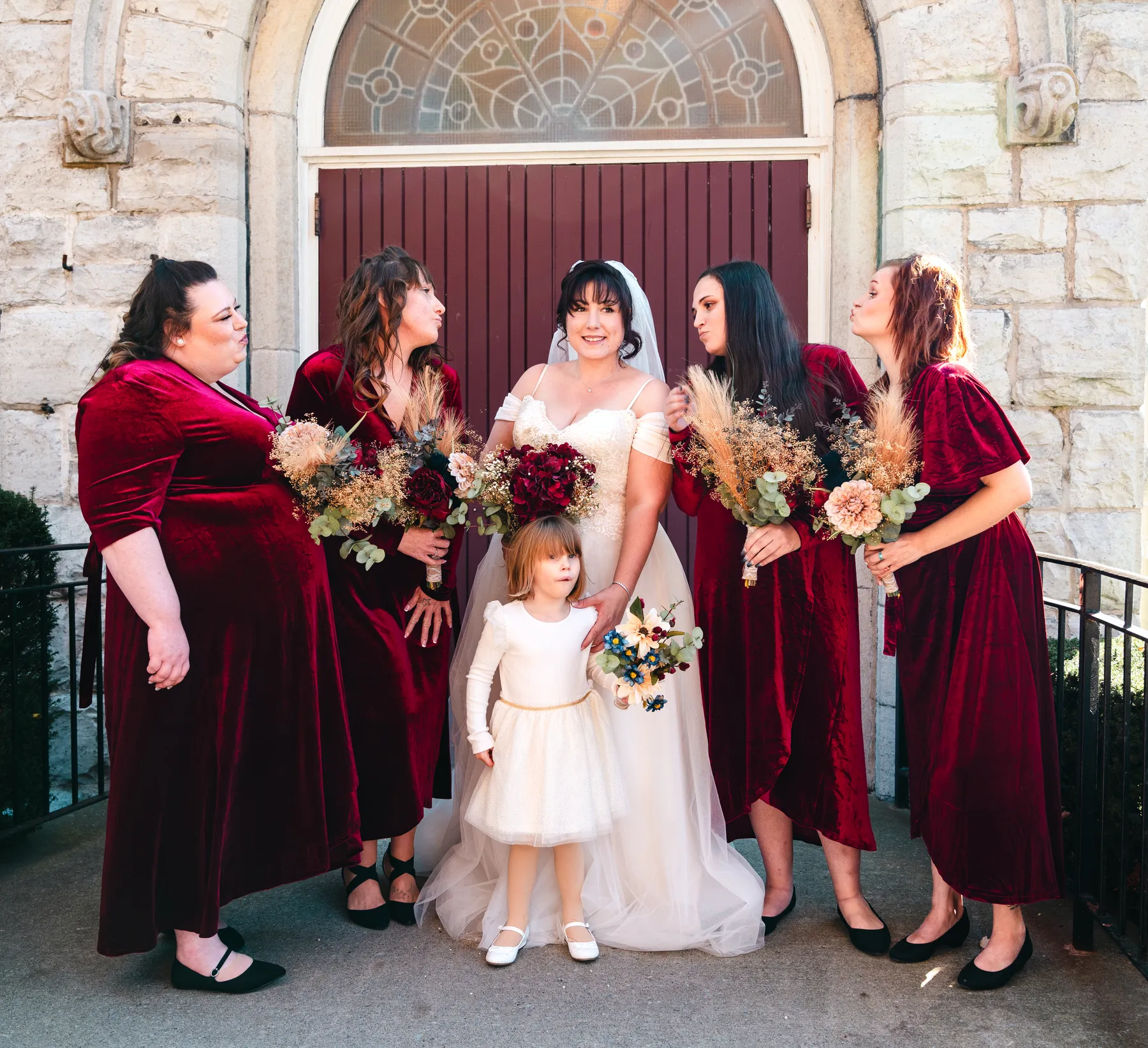 The bride with bridesmaids and a flower girl at the church door at Kody and Jaz’s wedding.