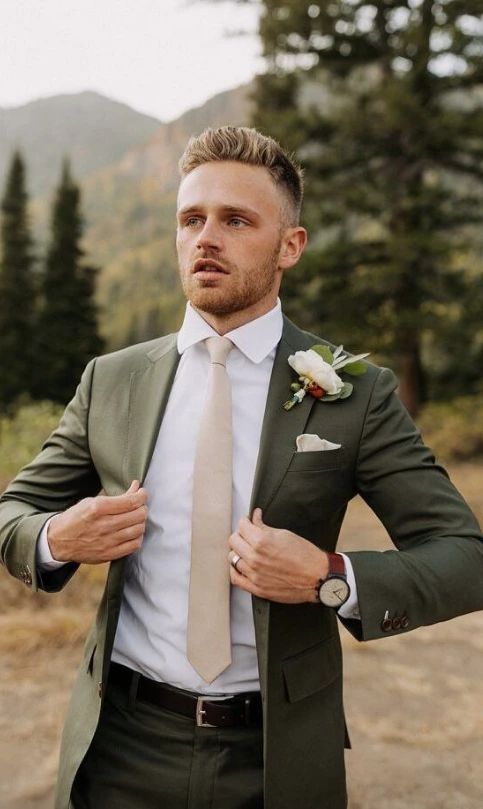 A groom wearing a forest green suit, white shirt, beige tie, and boutonniere, standing outdoors with mountains and trees in the background.