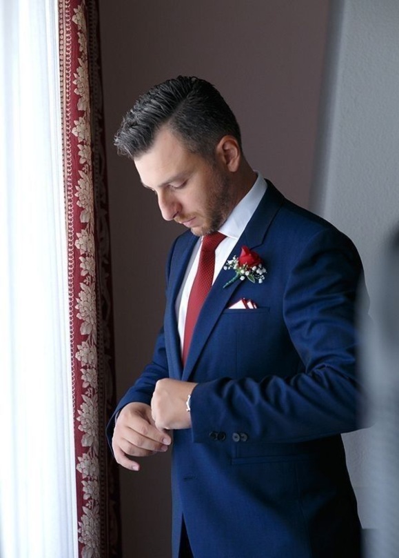 A groom in a navy suit with a burgundy tie and boutonniere, standing near a window with red curtains.