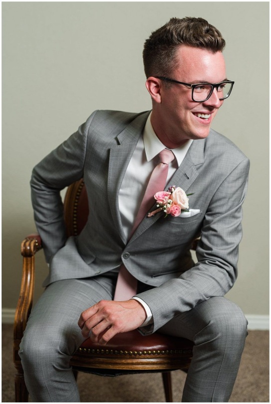 A groom wearing a charcoal gray suit, white shirt, pink tie, and boutonniere, sitting on a wooden chair with a floral pocket square.