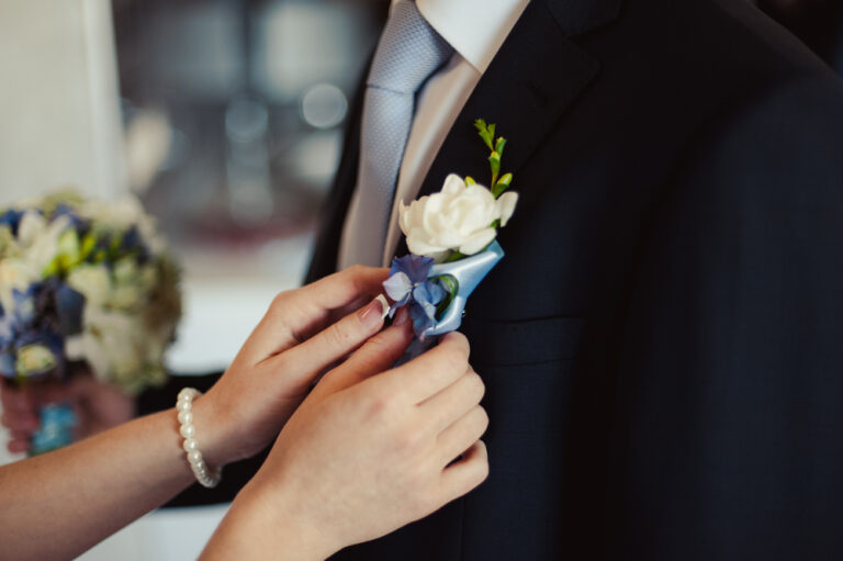 Groom suit accessories with light blue tie and white floral boutonniere being pinned.
