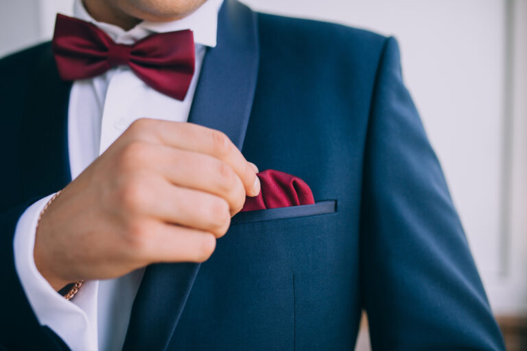 Groom suit accessories with burgundy bow tie and matching pocket square in navy suit.